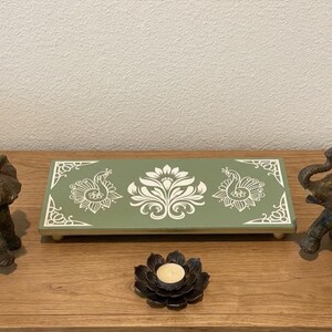 May include: A decorative display featuring two elephant figurines flanking a green tray with a white floral design and peacock motifs. A black lotus-shaped candle holder with a lit candle sits in front.