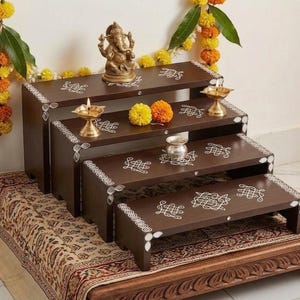 May include: A four-tiered dark brown wooden display stand featuring white painted geometric patterns on the edges and surfaces. The stand is arranged in a cascading staircase formation, holding a brass Ganesha figurine, two brass oil lamps, and a small silver vessel.