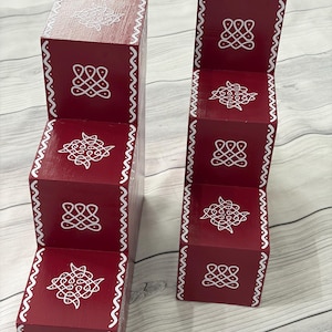May include: Two red wooden step-shaped decorative blocks. Each step is adorned with white, intricate patterns and a wavy border. The blocks are designed with a stair-like structure, showcasing a unique and artistic design.