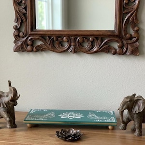 May include: A wooden table displays decorative items, including two bronze elephant figurines, a teal tray with a lotus design, and a bronze lotus flower. A carved wooden mirror hangs above the table.