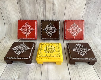 May include: Five wooden square platforms with raised edges, painted in different colors and decorated with white intricate patterns. The platforms are arranged in a row on a woodgrain surface.