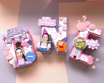 Puede incluir: Letras 3D rosas y moradas que deletrean "ONE" con un tema de chicle. Las letras están decoradas con lunares rosas y blancos, burbujas y una máquina de chicles. La letra "N" tiene una imagen de una persona con un gorro de fiesta y un monstruo naranja peludo. La letra "E" tiene una máquina de chicles con chicles de colores y un bocadillo que dice "¡Pop!" La letra "O" tiene una imagen de una persona con el texto "Can you say one?" encima de la imagen.