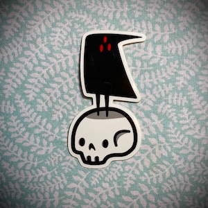 Könnte beinhalten: Ein Aufkleber mit einem schwarzen Raben mit roten Augen, der auf einem weißen Schädel sitzt.