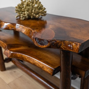 May include: A two-tiered wooden console table with a live edge top and bottom shelf. The table is made of dark brown wood with a natural finish. The table is supported by four sturdy legs.