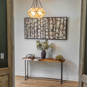 Puede incluir: Una mesa consola de madera con una tapa de borde vivo y patas de metal negro. La mesa est&aacute; decorada con un jarr&oacute;n de flores y una pieza decorativa. Un cuadro abstracto enmarcado cuelga en la pared detr&aacute;s de la mesa.