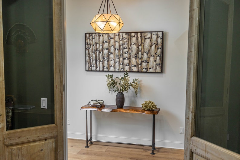 Puede incluir: Una mesa consola de madera con una tapa de borde vivo y patas de metal negro. La mesa est&aacute; decorada con un jarr&oacute;n de flores y una pieza decorativa. Un cuadro abstracto enmarcado cuelga en la pared detr&aacute;s de la mesa.