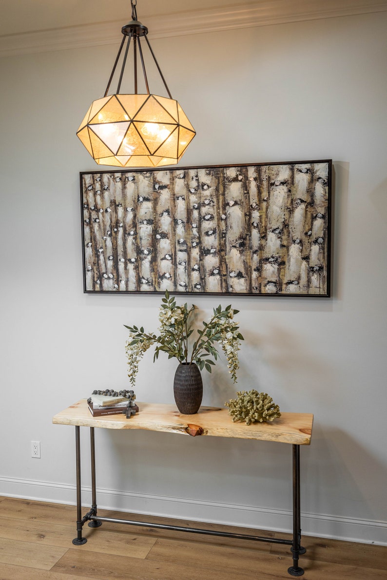 Puede incluir: Una mesa consola de madera con patas de tubo de metal negro. La mesa tiene una tapa de borde vivo y est&aacute; decorada con un jarr&oacute;n de flores blancas, una pila de libros y un trozo de coral. Una l&aacute;mpara geom&eacute;trica cuelga sobre la mesa.