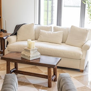 May include: A white linen sofa with pillows and a throw blanket sits in a living room with a large window.  The sofa is in front of a coffee table made of wood with a live edge.  There are two side tables made of wood with live edges on either side of the sofa.