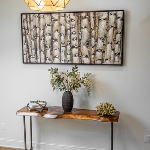 Puede incluir: Una mesa consola de madera con patas de tubo de metal negro. La mesa tiene una tapa de borde vivo y est&aacute; decorada con un jarr&oacute;n de flores, una pila de libros y un collar de cuentas. Un gran cuadro abstracto cuelga en la pared detr&aacute;s de la mesa.