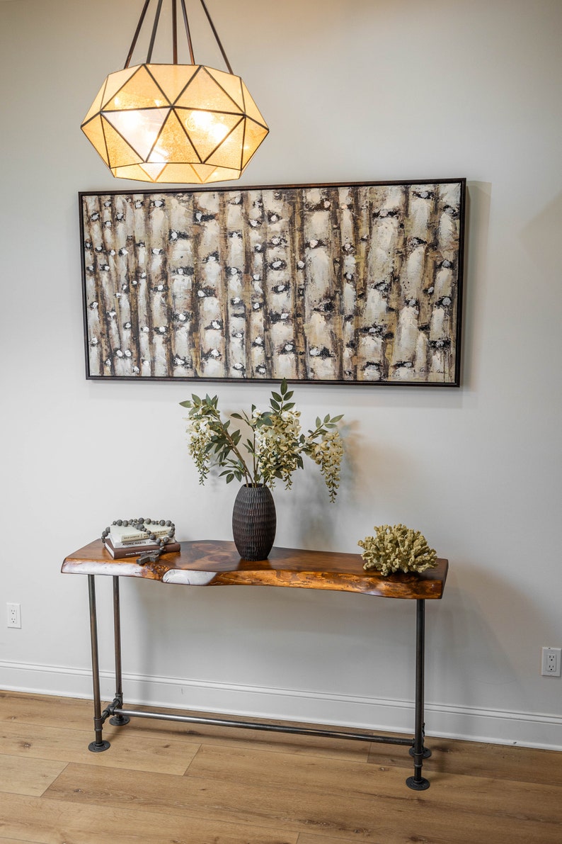 Puede incluir: Una mesa consola de madera con patas de tubo de metal negro. La mesa tiene una tapa de borde vivo y est&aacute; decorada con un jarr&oacute;n de flores, una pila de libros y un collar de cuentas. Un gran cuadro abstracto cuelga en la pared detr&aacute;s de la mesa.