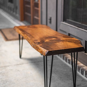 May include: A wooden bench with a live edge top and black hairpin legs. The bench is made of natural wood and has a rustic look.