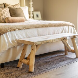 Pode incluir: Um banco de madeira clara com um aspecto natural e inacabado. O banco tem um assento retangular e pernas anguladas, colocado aos p&eacute;s de uma cama com roupa de cama em tons neutros. A sala tem uma luz suave e natural.