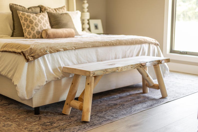 Pode incluir: Um banco de madeira clara com um aspecto natural e inacabado. O banco tem um assento retangular e pernas anguladas, colocado aos p&eacute;s de uma cama com roupa de cama em tons neutros. A sala tem uma luz suave e natural.