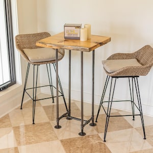 May include: A wooden table with a live edge top and black metal legs. Two wicker chairs with beige cushions are placed around the table. The table is decorated with a small box and two candles.