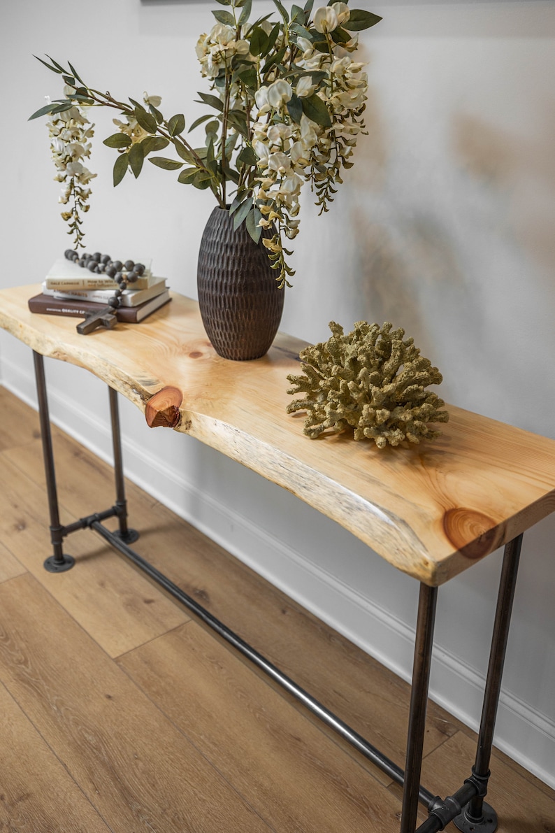 Puede incluir: Una mesa consola de madera con una tapa de borde vivo y patas de tubo de metal negro. La mesa est&aacute; decorada con un jarr&oacute;n de flores blancas y una pieza de coral.