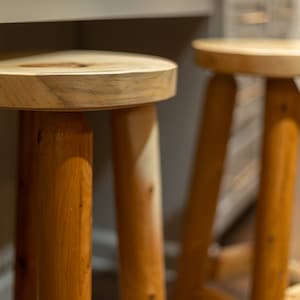 May include: Two wooden stools with round seats and three legs each. The stools are made of light-colored wood, with visible wood grain. The legs are cylindrical and the seats are flat. The stools are in a kitchen or dining setting.