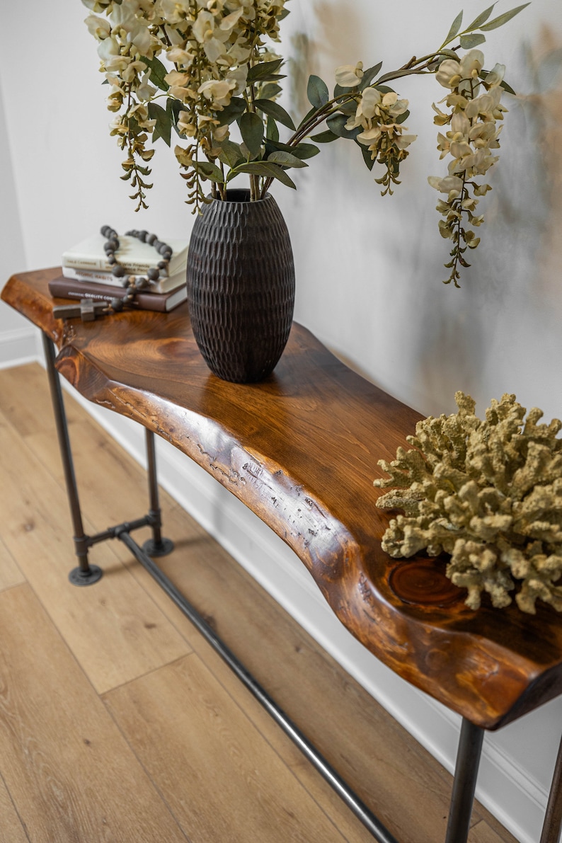 Puede incluir: Una mesa consola de madera r&uacute;stica con una tapa de borde vivo y patas de tubo de metal negro. Un jarr&oacute;n negro con flores blancas y una pieza decorativa de coral est&aacute;n sobre la mesa.