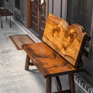 May include: A rustic wooden bench with a natural wood finish and a live edge design. The bench has a back and is made of solid wood. The bench is sitting on a concrete porch.