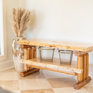 May include: A rustic wooden bench with two shelves, made from natural wood with a live edge. The bench has a natural wood finish and is supported by four sturdy legs. Two wire baskets are on the shelves.