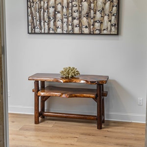 Puede incluir: Una mesa consola de madera rústica con una tapa de borde vivo y dos estantes. La mesa está hecha de madera marrón oscura y tiene un acabado natural. Un pequeño trozo de coral decorativo se encuentra en el estante superior.