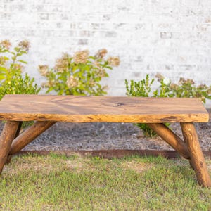 Pode incluir: Banqueta de madeira r&uacute;stica com acabamento natural. A banqueta tem um tampo retangular e pernas anguladas em estilo tronco. Est&aacute; sobre um leito de relva verde com uma parede de tijolos brancos ao fundo. A banqueta tem cerca de 122 cm de comprimento.