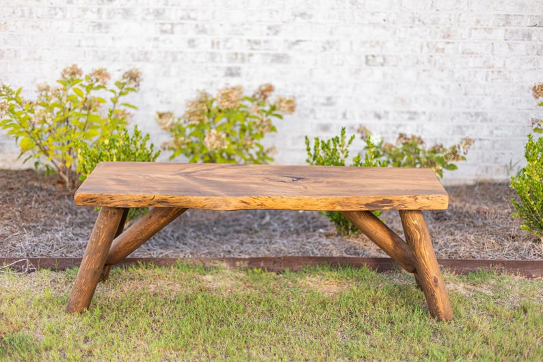 Pode incluir: Banqueta de madeira r&uacute;stica com acabamento natural. A banqueta tem um tampo retangular e pernas anguladas em estilo tronco. Est&aacute; sobre um leito de relva verde com uma parede de tijolos brancos ao fundo. A banqueta tem cerca de 122 cm de comprimento.
