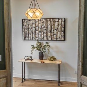 Puede incluir: Una mesa consola de madera con una tapa de borde vivo y patas de metal negro. Un jarr&oacute;n con flores blancas y un objeto decorativo est&aacute;n sobre la mesa. Una l&aacute;mpara geom&eacute;trica cuelga del techo sobre la mesa.