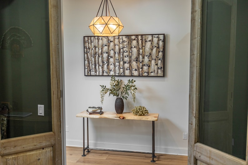 Puede incluir: Una mesa consola de madera con una tapa de borde vivo y patas de metal negro. Un jarr&oacute;n con flores blancas y un objeto decorativo est&aacute;n sobre la mesa. Una l&aacute;mpara geom&eacute;trica cuelga del techo sobre la mesa.