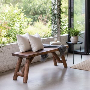 Pode incluir: Banqueta de madeira r&uacute;stica com acabamento natural, com duas almofadas brancas. Uma manta cinza e livros repousam na banqueta. A banqueta est&aacute; situada num p&aacute;tio de cimento, com vegeta&ccedil;&atilde;o ao fundo.