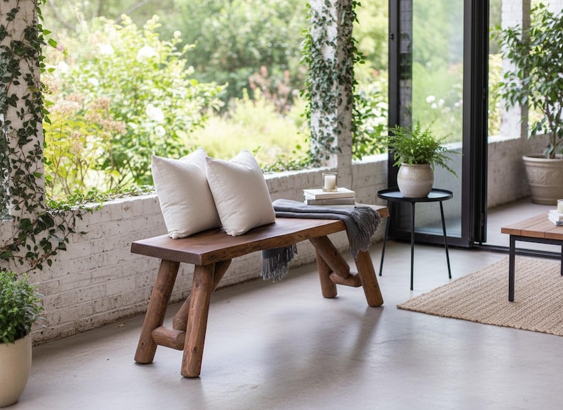 Pode incluir: Banqueta de madeira r&uacute;stica com acabamento natural, com duas almofadas brancas. Uma manta cinza e livros repousam na banqueta. A banqueta est&aacute; situada num p&aacute;tio de cimento, com vegeta&ccedil;&atilde;o ao fundo.