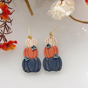 Puede incluir: Un par de pendientes colgantes con un diseño apilado de tres calabazas en color crema, naranja y azul oscuro con postes dorados. Las calabazas azul oscuro tienen motas doradas y pequeñas flores azules adheridas a los lados de las calabazas.