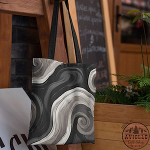 Op de afbeelding: Een zwart-witte tote bag met een draaiend abstract patroon. De tas heeft zwarte banden en een logo met de tekst "Aviellie Gift Shop" met een dennenboomontwerp.