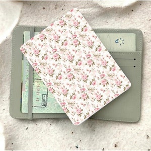 May include: A grey passport holder with a white floral pattern featuring pink roses. The holder has a flap that closes with a snap.