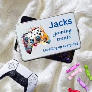 May include: A white tin with a colourful illustration of a video game controller. The tin has the text "Jacks gaming treats Leveling up every day" printed on it. There are also several colourful wrapped sweets on a white fluffy surface.