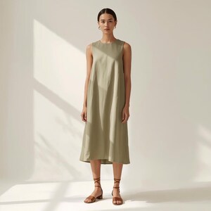 Puede incluir: Vestido midi verde oliva sin mangas con cuello redondo. El vestido se combina con sandalias de cuero marr&oacute;n. La modelo est&aacute; de pie frente a una pared blanca con sombras.