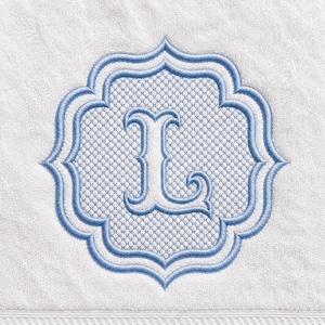 Puede incluir: Una toalla de felpa blanca con un diseño bordado centrado. El diseño consiste en un marco de cuatrifolio azul claro con un patrón de celosía blanco en el interior, que rodea una letra L mayúscula estilizada en hilo azul claro.
