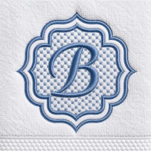 May include: A white terry cloth towel featuring an embroidered quatrefoil frame in blue and white. The center of the frame contains a repeating small blue star pattern and a large, prominent blue embroidered letter B in a cursive script font.