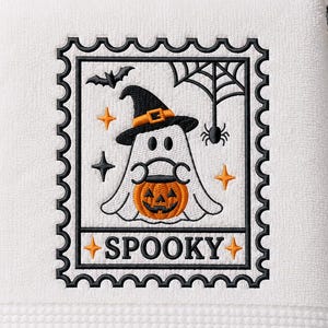May include: A white hand towel featuring an embroidered postage stamp design. The embroidery depicts a ghost wearing a black witch hat and holding an orange jack-o'-lantern, surrounded by a bat, spider web, and stars, with the word SPOOKY below.