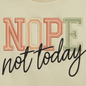 以下が含まれることがあります： 薄いベージュのトレーナーに「NOPE not today」という文字がピンク、オレンジ、グリーンで刺繍されています。