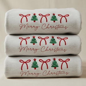 May include: Three folded light grey towels stacked. Each towel is embroidered with red bows, green Christmas trees, and the words "Merry Christmas" in red script.