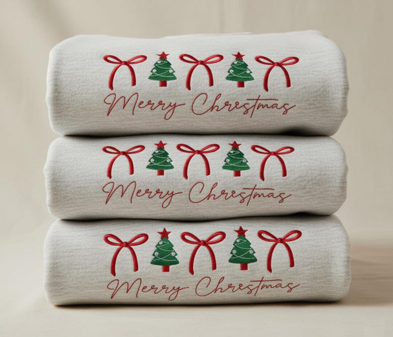 May include: Three folded light grey towels stacked. Each towel is embroidered with red bows, green Christmas trees, and the words "Merry Christmas" in red script.