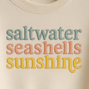 以下が含まれることがあります： パステルカラーの刺繍が施されたクリーム色のスウェットシャツ。「saltwater seashells sunshine」の文字が重ねて配置されています。刺繍は青、桃色、黄色の色合いです。