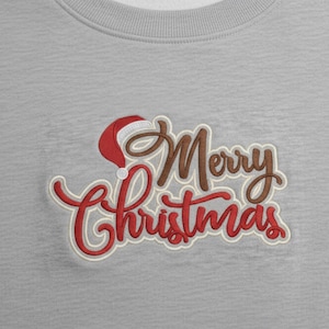 Puede incluir: Sudadera gris claro con la frase bordada "Merry Christmas" en escritura roja y marr&oacute;n. Un gorro de Pap&aacute; Noel est&aacute; incorporado en el dise&ntilde;o, a&ntilde;adiendo un toque festivo a la ropa navide&ntilde;a.