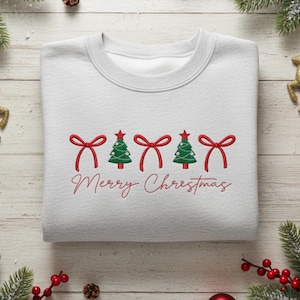 May include: A white sweatshirt featuring a Christmas design. The design includes red bows, green Christmas trees, and the words "Merry Christmas" in red script. The sweatshirt is neatly folded.