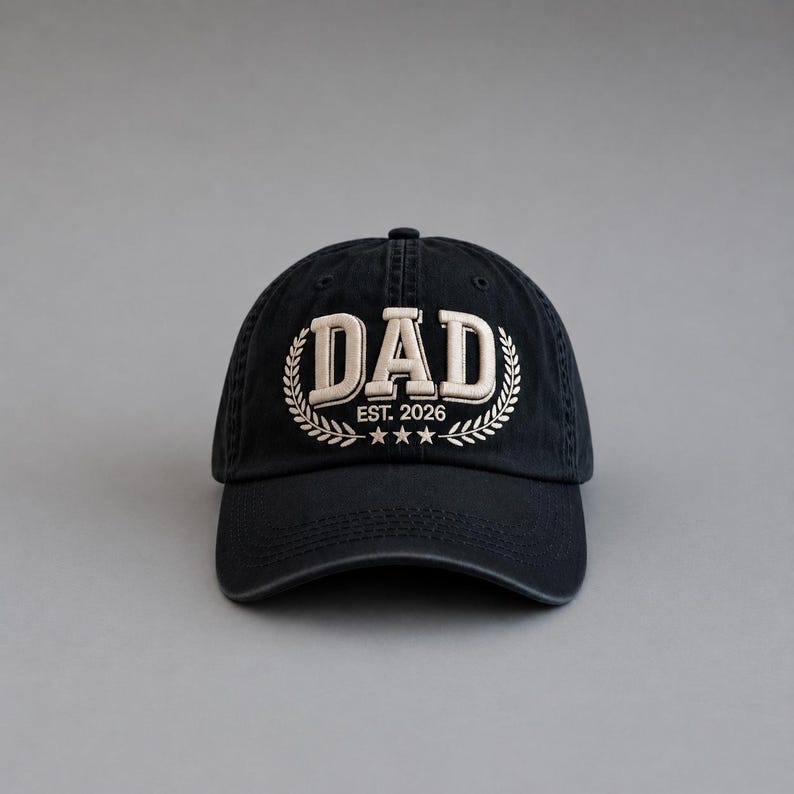 May include: Black baseball cap with the word "DAD" embroidered in white, surrounded by a laurel wreath design. The text "EST. 2026" and three stars are below the word "DAD".
