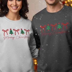 May include: Two sweatshirts, one light grey and one charcoal, feature a festive design. Each has red bows, green Christmas trees, and the words "Merry Christmas" in a script font. Ideal for the holiday season.