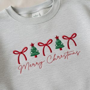 May include: Pale grey sweatshirt featuring red embroidered bows, green Christmas trees, and the words "Merry Christmas" in red script. The trees are topped with red stars.