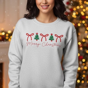 May include: A light grey sweatshirt featuring a Christmas design. The design includes red bows, green Christmas trees, and the words "Merry Christmas" in a cursive font. The sweatshirt is a long-sleeved crew neck.