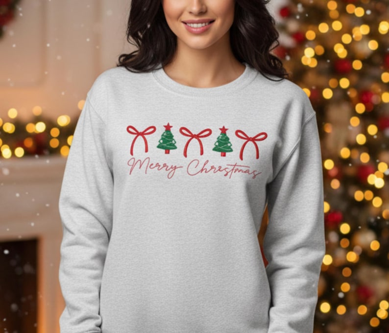 May include: A light grey sweatshirt featuring a Christmas design. The design includes red bows, green Christmas trees, and the words "Merry Christmas" in a cursive font. The sweatshirt is a long-sleeved crew neck.