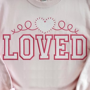 May include: Pink sweatshirt with a pink embroidered heart and the word "LOVED" in white letters with a pink outline.